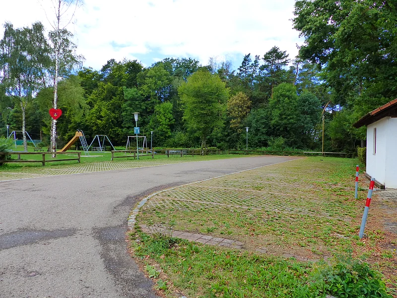 Parkplatz mit Grünfläche rechts im Bild und einem Spielplatz mit Rutsche und Schaukeln vor Wald im Hintergrund. Ein Maibaum mit Herz steht auf der Grünfläche links.
