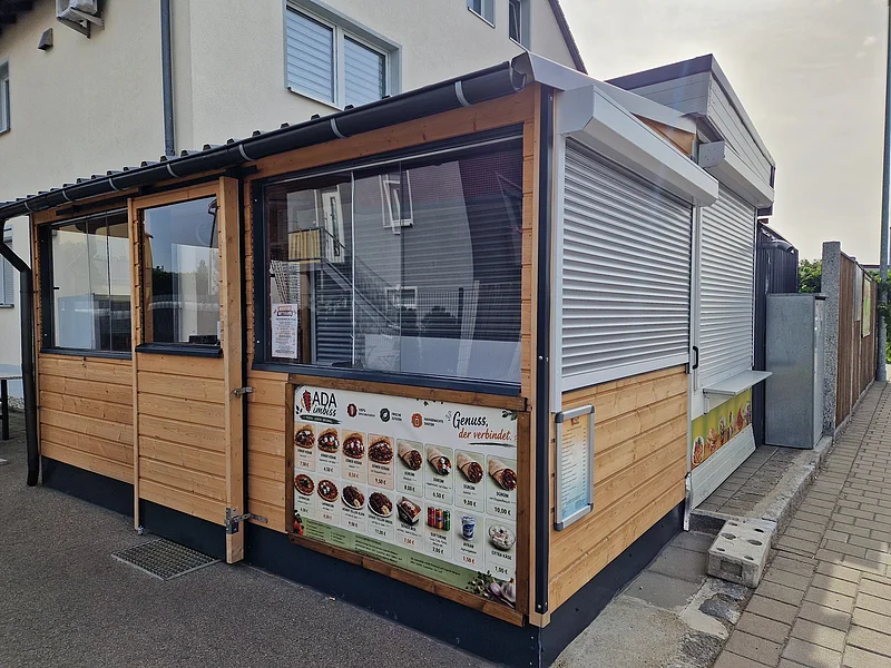 Imbiss ADA Döner-Kebap in Monheim an der Wemdinger Straße Holzverkleideter Döner-Imbissstand mit teilweise geschlossenen Rollläden und einer Speisekarte an der Außenwand rechts unten, Gehweg daneben.