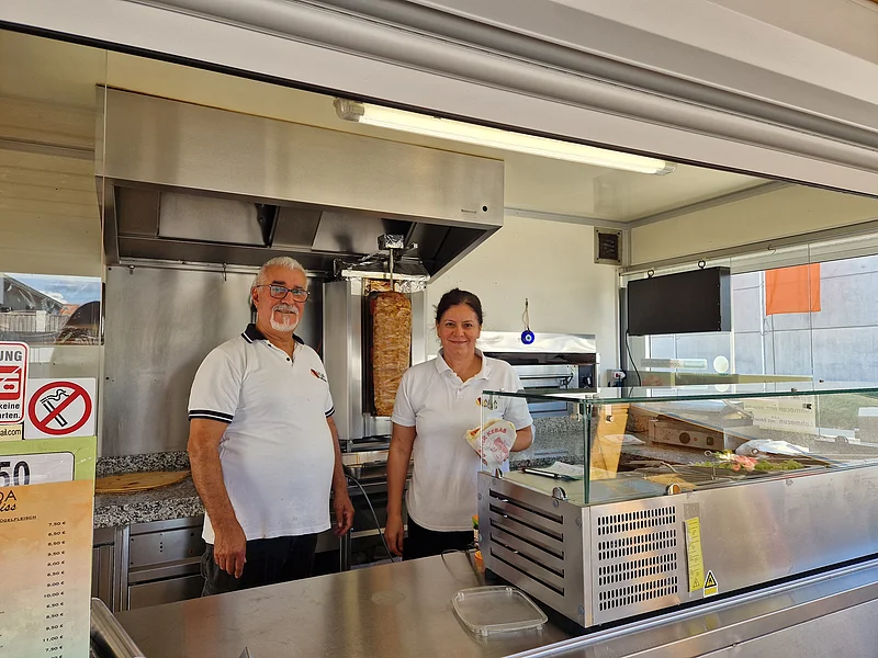 Inhaber vom "Ada Imbiss Döner Kebab" Monheim Zwei Personen in weißen Poloshirts stehen in einem Imbiss mit Dönergrill und Theke und blicken lächelnd in die Kamera.