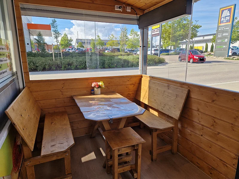 Sitzmöglichkeit beim "Ada Imbiss Döner Kebab" in Monheim Holzsitzecke mit Tisch in einem Imbiss mit Blumen auf dem Tisch. Durch das Fenster Blick auf Parkplatz und Aldi-Süd-Schild bei Tageslicht