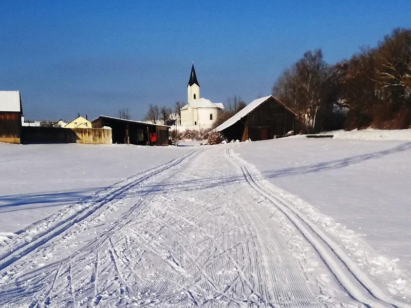 Verschneite Landschaft mit Skispuren, ein Ort zum Langlaufen in der Monheimer Alb. Im Hintergrund Gebäude, eine Kirche und blauer Himmel zu sehen.