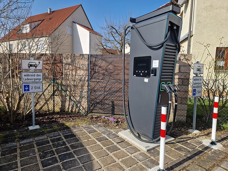 Öffentlich zugängliche Ladesäule an der Raiffeisen-Volksbank Monheim Schwarze Elektroauto-Ladestation mit zwei Ladepunkten stehend vor einem Parkplatz mit Schildern zur Parkzeitbegrenzung auf 2 Stunden, bei Tageslicht. Im Hintergrund Wohnhäuser vor blauem Himmel.