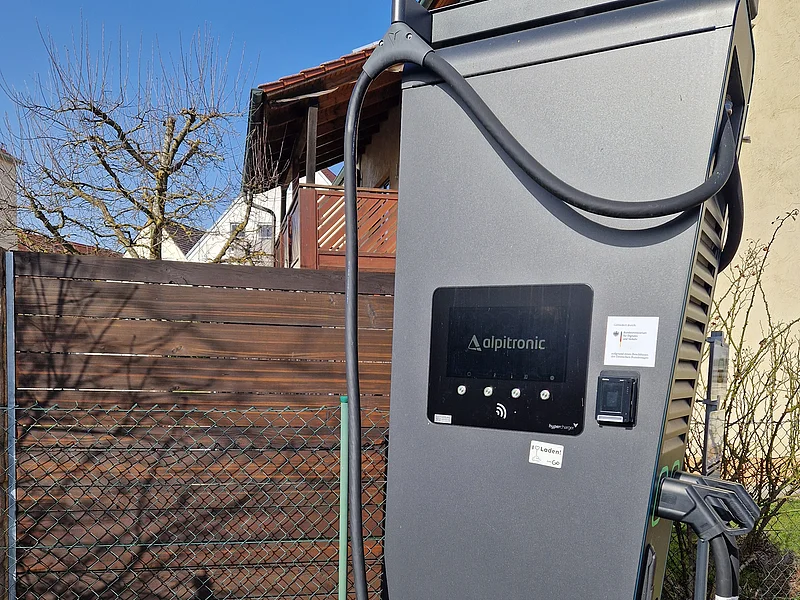 Ladesäule an der Raiffeisen-Volksbank Monheim Schwarze Elektroauto-Ladesäule der Marke Alpitronic mit einem Display und zwei Schläuchen zum Laden, vor einem Holzzaun und einem Wohnhaus im Hintergrund, bei Sonnenschein.