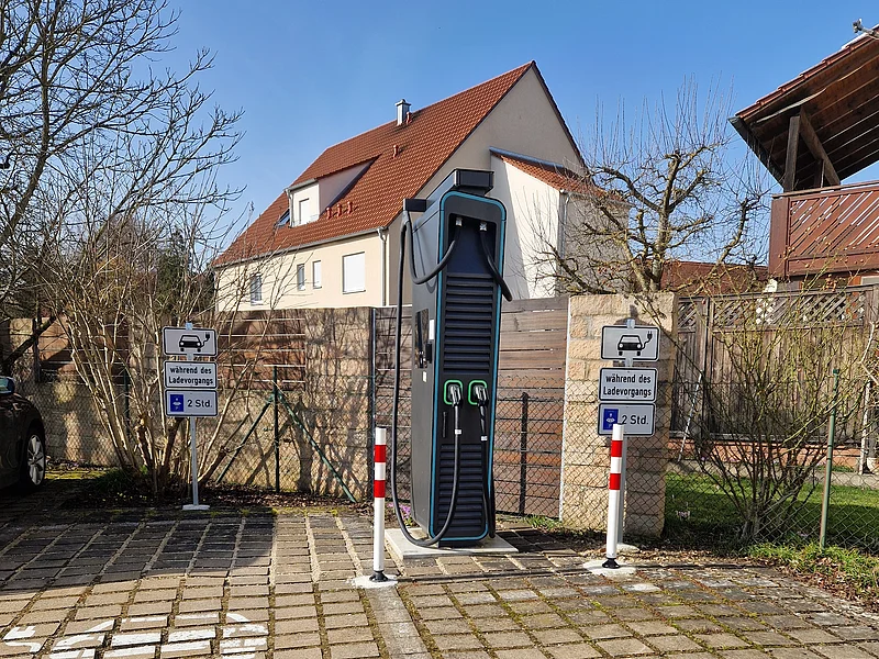 Zwei Parkplätze vor der Ladesäule an der Raiffeisen-Volksbank Monheim Schwarze Elektroauto-Ladestation mit zwei Ladepunkten hinter zwei gepflasterten Parkplätzen vor einem Wohnhaus bei klarem Himmel.