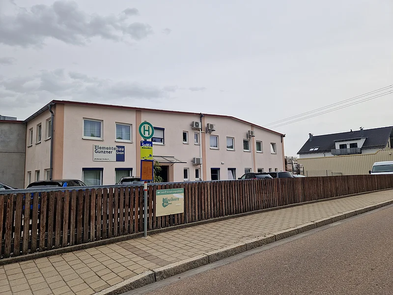 Elementbau Gunzner in Monheim Zweistöckiges Bürogebäude mit Parkplätzen hinter Holzzaun und Bushaltestelle im Vordergrund bei bewölktem Himmel.