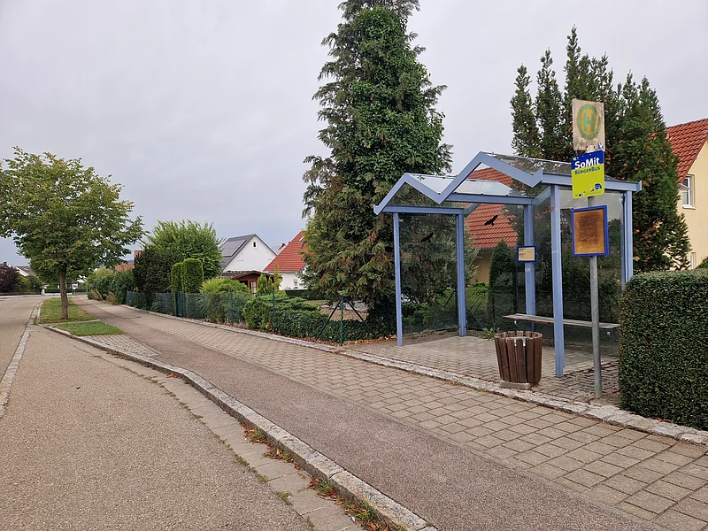Haltestelle mit Wartehäuschen und Mülleimer an einer ruhigen Straße mit Bäumen und Wohnhäusern im Hintergrund.