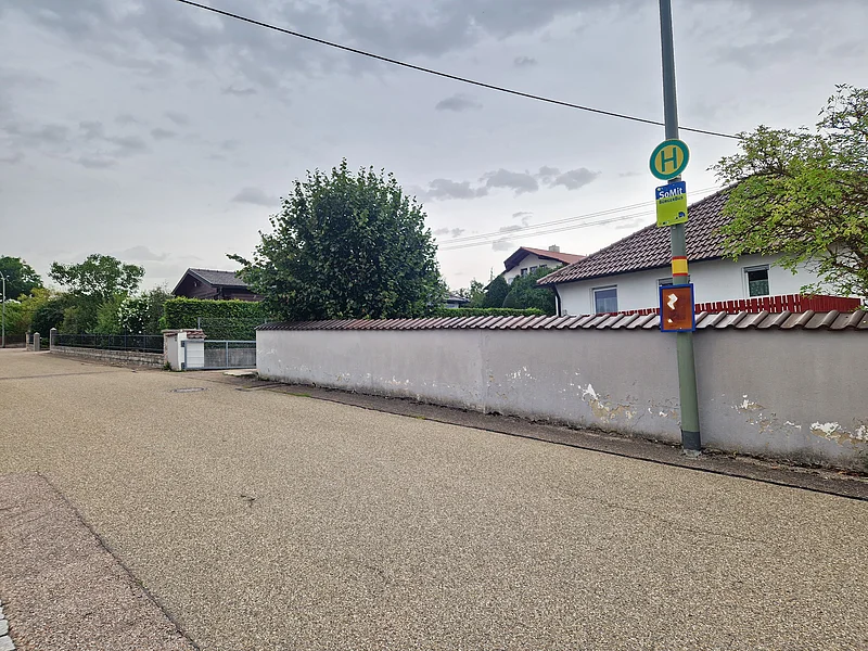 Straße im Wohngebiet mit Bushaltestelle vor Haus mit Ziegeldach und Bäumen, bei bewölktem Himmel.