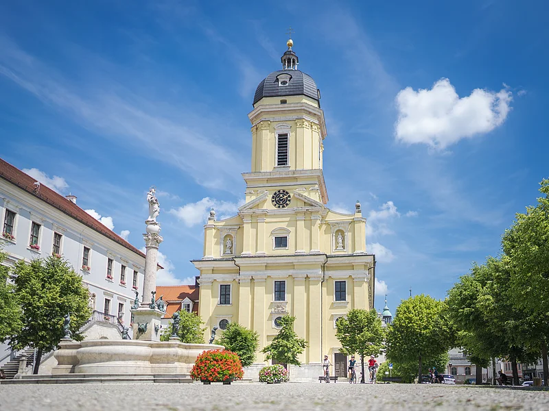 Radelnde auf dem Karlsplatz vor der prächtigen Hofkirche. Im Vordergrund ist ein Brunnen zu sehen.