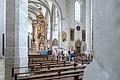 Zwischen zwei Säulen hindurch geht der Blick auf eine Gruppe, die sich in einem Gang zwischen Kirchenbänken auf einen Altar zubewegt.