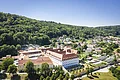 Luftaufnahme eines großen Gebäudekomplexes mit roten Dächern vor bewaldeten Hügeln und einem Dorf im Hintergrund.
