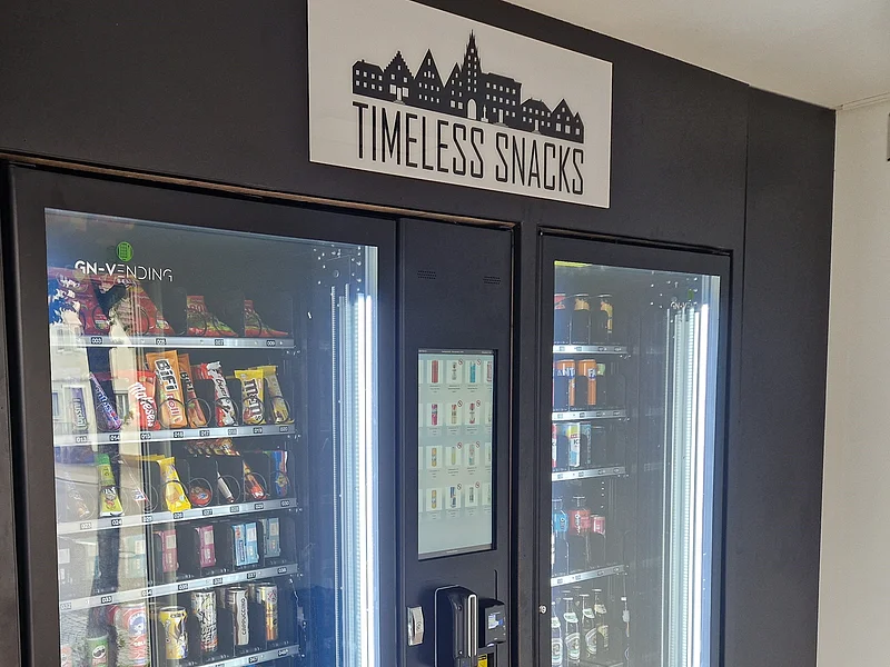 Zwei schwarze Snack- und Getränkeautomaten mit beleuchteten Sichtfenstern und Bedienfeld, darüber Schild „TIMELESS SNACKS“.