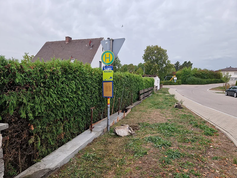 Bushaltestelle mit grünem Heckenzaun, Gehweg und Wohnhäusern bei bewölktem Himmel