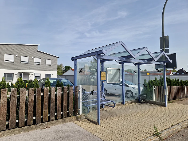 Haltestelle an einem Gehweg mit blauer Sitzbank und Glaswänden vor Wohnhäusern und geparkten Autos im Hintergrund, bei klarem Himmel.