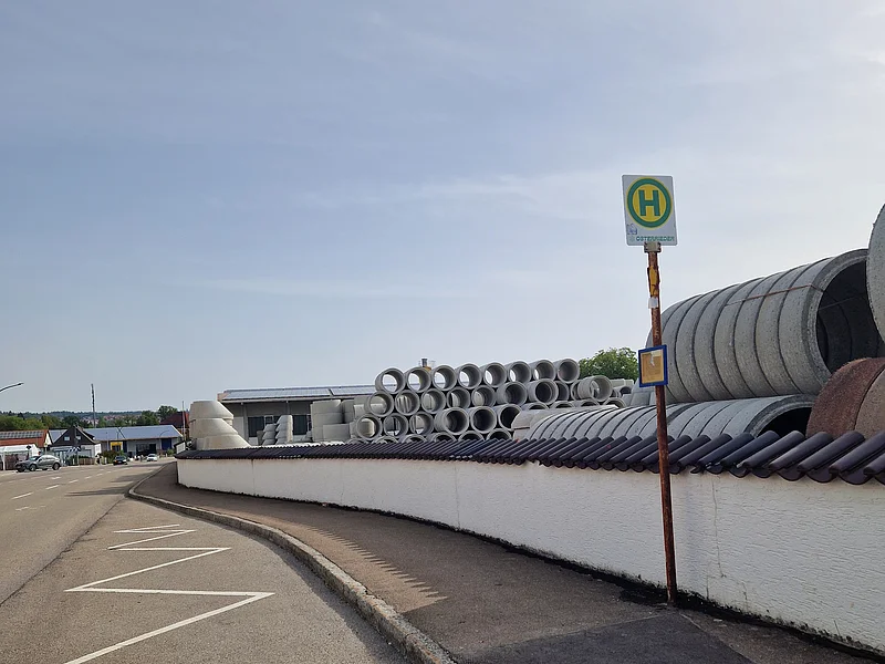 Einbuchtung einer Bushaltestelle mit dazugehörigem Schild am Gehweg neben Lagerplatz mit Betonrohren hinter weißer Mauer, bei klarem Himmel