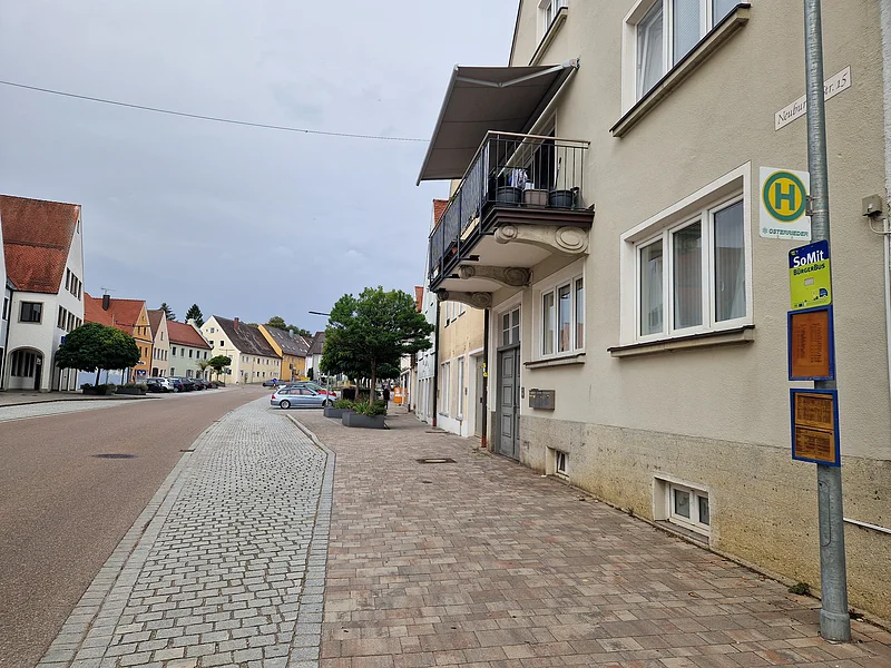Straße mit Bürgersteig, Häusern, Bäumen und Bushaltestelle mit Fahrplänen an Laterne bei bewölktem Himmel