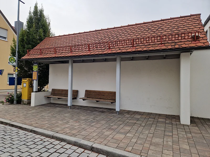 Überdachte Bushaltestelle mit zwei Holzbänken und rotem Ziegeldach an gepflastertem Gehweg.