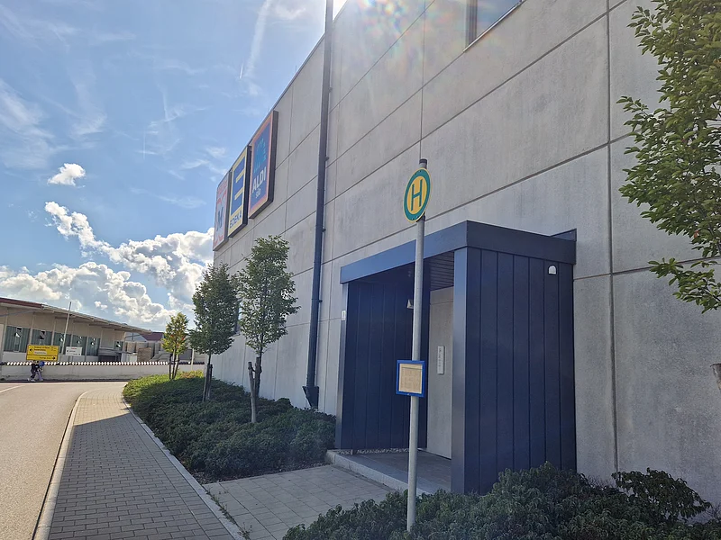Haltestelle mit Schild vor einer grauen Gebäudewand mit ALDI- und EDEKA-Schildern unter blauem Himmel mit Wolken. Links im Bild eine Verkehrsstraße.