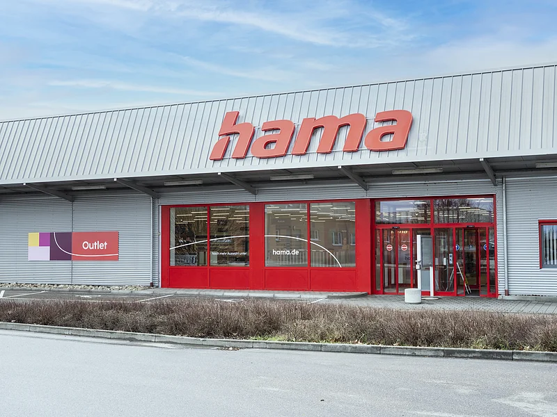 Hama Outlet Graue Fassade eines Outlet-Geschäfts mit rotem Schriftzug "Hama", roten Eingangstüren und roten Fensterumrandungen, bei bewölktem Himmel. Vor dem Geschäft eine Hecke entlang einer Straße.