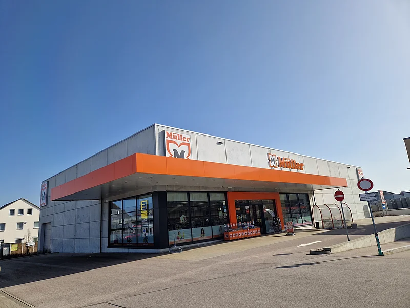 Drogeriemarkt Müller - Außenansicht der Filiale in Monheim Außenansicht eines Müller-Marktes mit orangefarbenem Vordach und klarem Himmel im Hintergrund auf einem gepflastertem Platz mit Parkplätzen außenrum.
