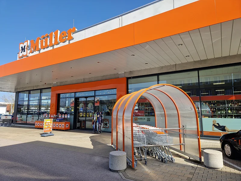 Drogeriemarkt Müller - Ansprechende Filiale in Monheim Eingangsbereich eines Müller-Marktes mit Einkaufswagen und orangen Markenzeichen "Müller" bei Sonnenschein.