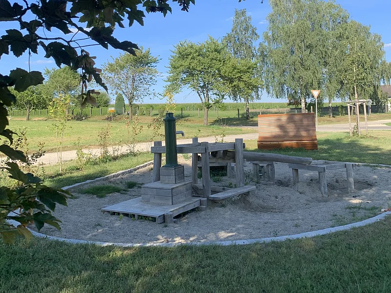 Dorferneuerung Flotzheim Wasserspielplatz Rund gestalteter Wasserspielplatz mit Holz-Wasserpumpe und Sandbereich, umgeben von einer Rasenfläche. Im Hintergrund Bäume und blauer Himmel.