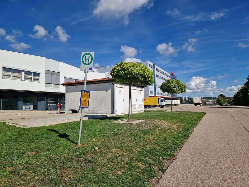 Haltestellenschild vor kleinem Gebäude mit zwei Bäumen und LKWs vor großem Industriegebäude unter blauem Himmel.