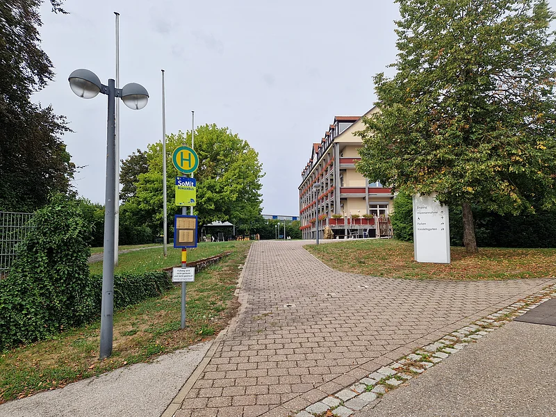 Buswartehäuschen mit Haltestellenschild an gepflastertem Weg vor mehrstöckigem Gebäude und Bäumen bei bewölktem Himmel