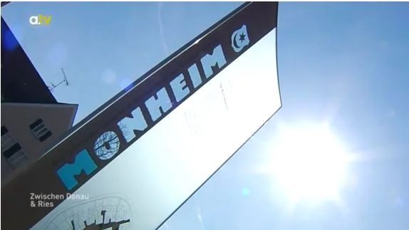 Ein Schild mit der Aufschrift "Monheim" vor einem blauen Himmel und Sonnenschein. Links oben das Logo von a.tv. Im Hintergrund ein Haus.