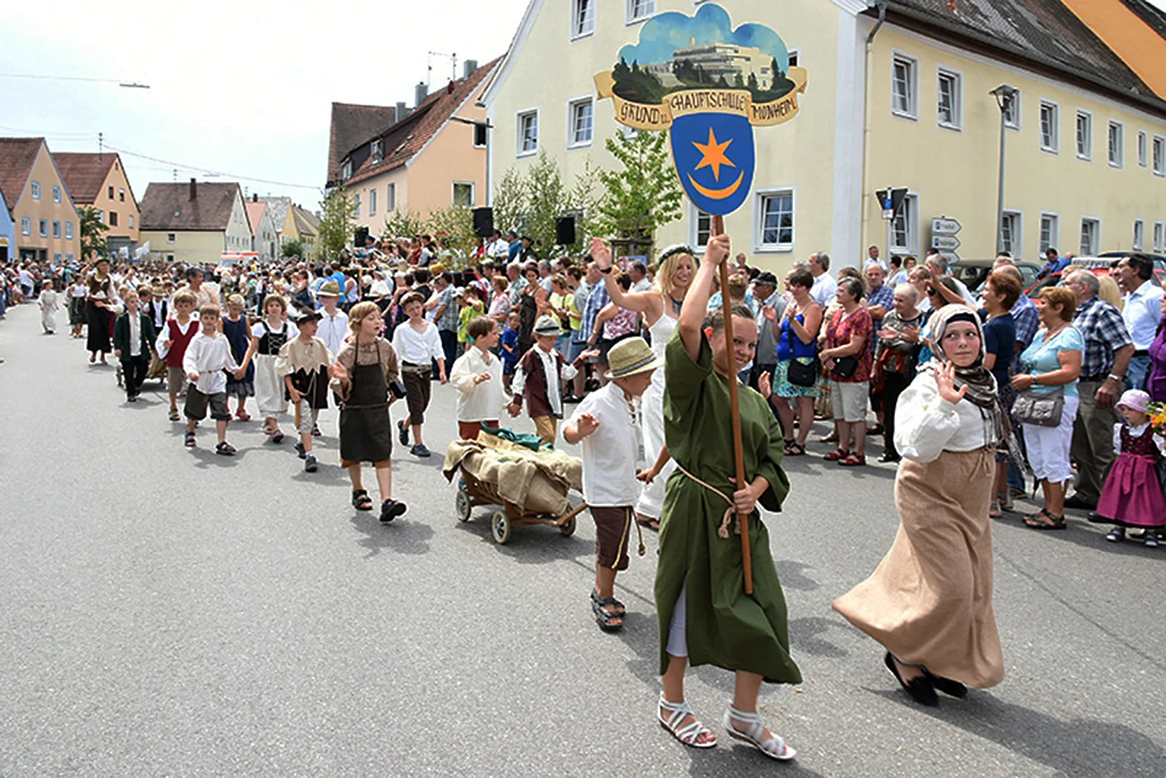 Ein historischer Festumzug bei dem größtenteils Kindern aber auch mit Erwachsenen in historischen Kleidern durch eine Straße mit vielen Zuschauer an den Häusern ziehen. Dabei hält das erste Kind einen Wappen in den Händen, andere ziehen einen kleinen Karren hinter sich her.