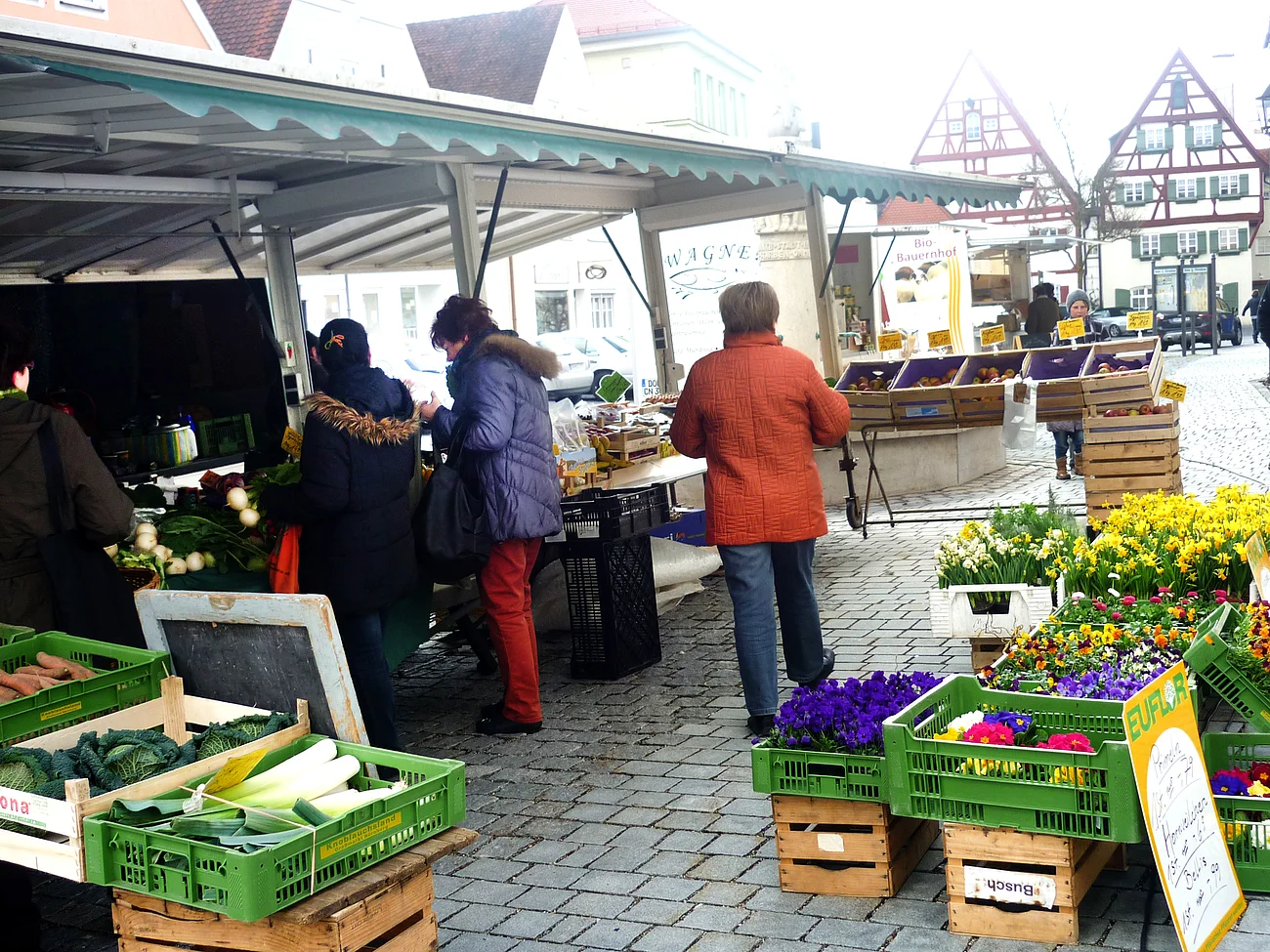 Menschen kaufen an einem Marktstand mit Gemüse und Blumen auf einem gepflasterten Platz ein.