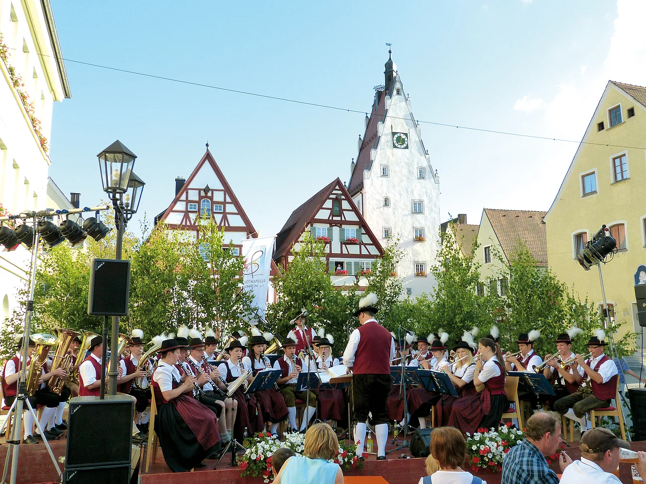 Blaskapelle in traditioneller Tracht spielt auf einer Bühne vor Fachwerkhäusern und Publikum bei Sonnenschein im Freien.