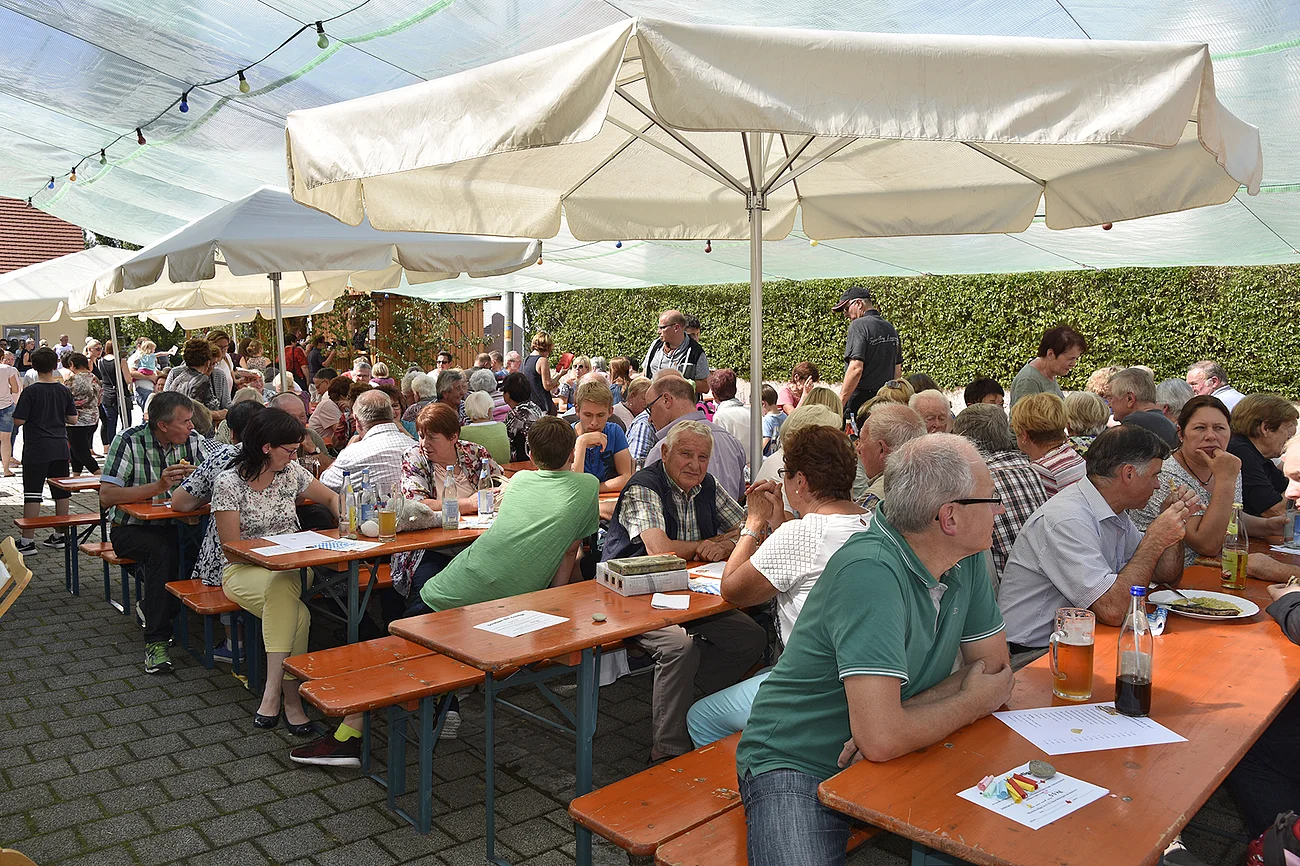 Viele Menschen sitzen an langen Holztischen unter weißen Sonnenschirmen im Freien bei einer Veranstaltung. Im Hintergrund eine hohe grüne Hecke.