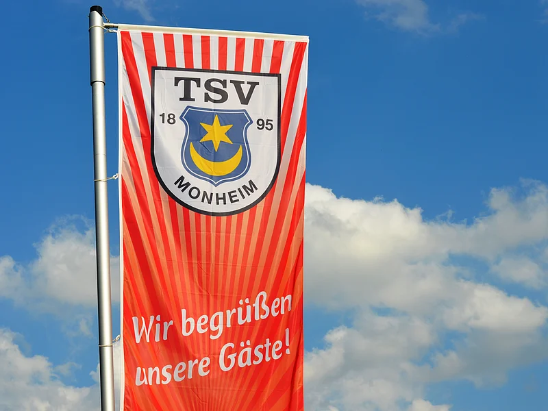 Rote Fahne mit dem Logo des TSV Monheim und dem Text "Wir begrüßen unsere Gäste!" vor blauem Himmel.