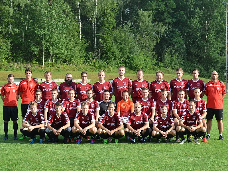 TSV 1895 Monheim e.V. - Sparte Fußball Fußballmannschaft mit 29 Spielern und Trainern in roten Trikots auf grünem Spielfeld vor Bäumen posierend.