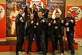 TSV 1895 Monheim e.V. - Sparte Karate Neun Personen in Sportkleidung posieren mit geballten Fäusten vor Werbebannern zum Karatesport und einem Sieger-Podest.