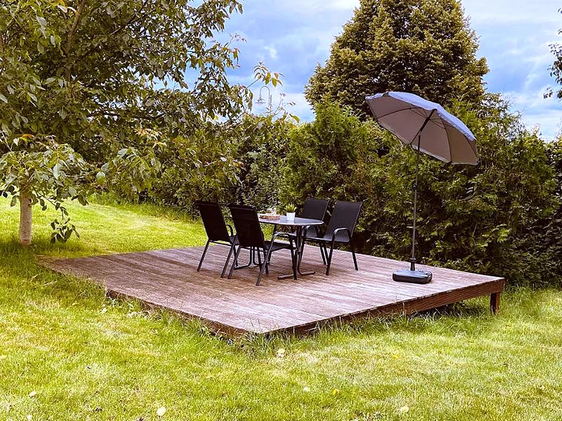 Holzterrasse mit Gartentisch, vier Stühlen und einem Sonnenschirm, umgeben von Rasen und Bäumen