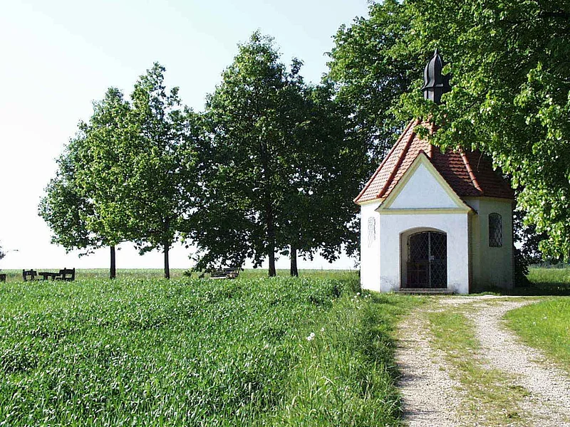 Kleine Kapelle mit rotem Dach auf einer Wiese mit einem Schotterweg. Umgeben von einem grünem Feld und Bäumen.