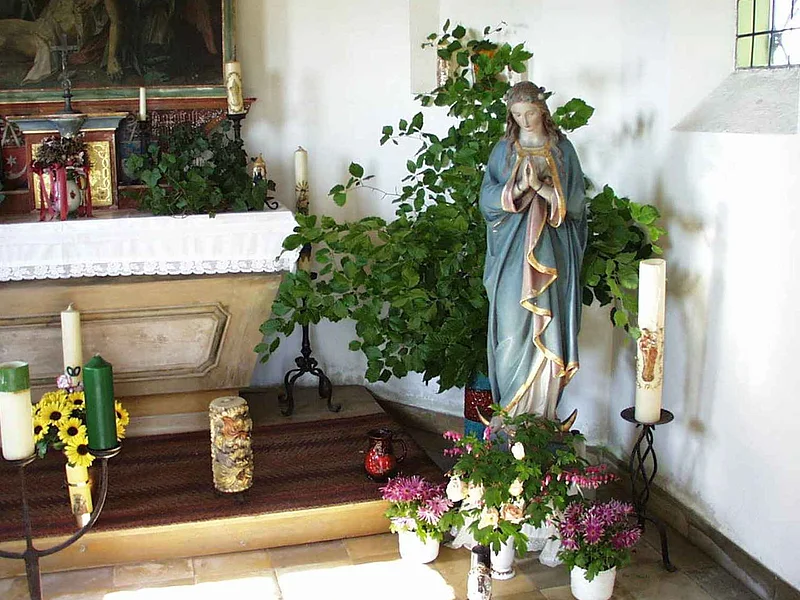 Innenansicht der Brandkapelle in Monheim Innenansicht einer kleinen Kapelle mit der Statue einer betenden Frau mit blauem Mantel in einer Ecke umgeben von Kerzen, Pflanzen und Blumen in einem weißen Raum. Links im Bild ein geschmückter Altar.