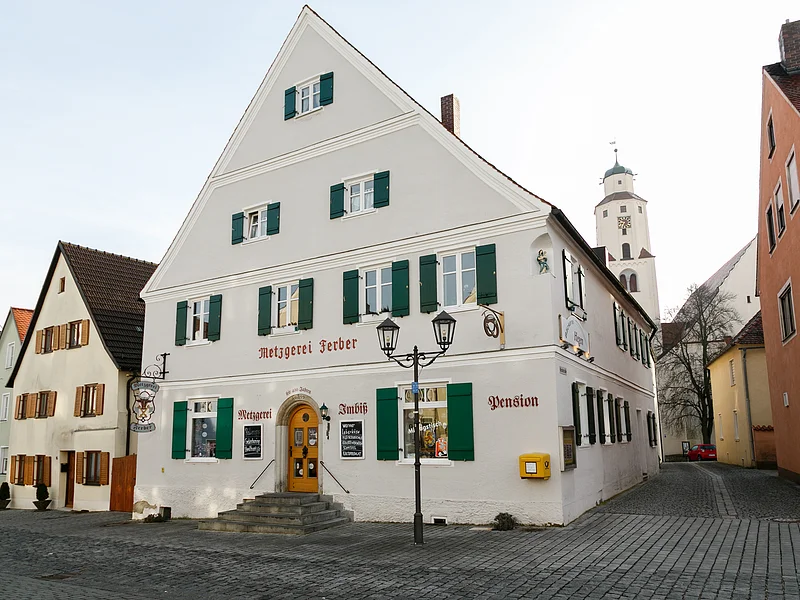 Graues mehrstöckiges Gebäude mit grünen Fensterläden, einer Treppe mit Geländern und dem Schriftzug "Metzgerei Ferber" oberhalb der Eingangstür, an einer gepflasterten Straße. Links von der Tür der Schriftzug "Metzgerei", rechts davon "Imbiss" und "Pension" an der Fassade. Ein gelber Briefkasten hängt an der vorderen Fassade.