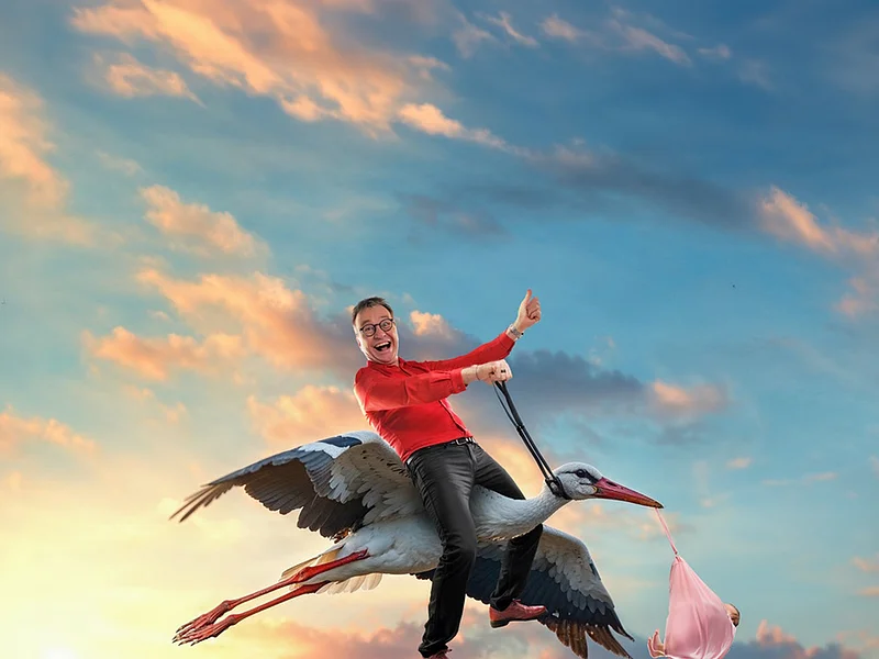 Pressebild Heinrich del Core Juhuu meine Frau wird Oma Mann in rotem Hemd reitet auf fliegendem Storch, der ein Baby in rosa Tuch trägt, vor Sonnenuntergangshimmel. Pressebild zur Veranstaltung Heinrich Del Core Juhuu meine Frau wird Oma am 09.04.2027 um 20.00 Uhr in der Stadthalle Monheim