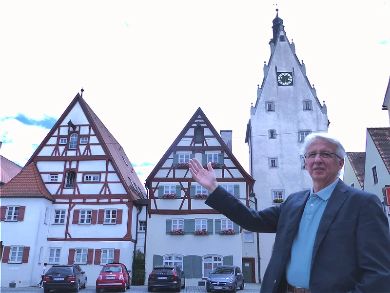 Eine Person mit Brille, im Sakko und blauem Hemd steht vor Fachwerkhäusern und einem Turm mit Uhr und zeigt darauf. Vor den Häusern stehen auf einer gepflasterten Fläche vier Autos geparkt.