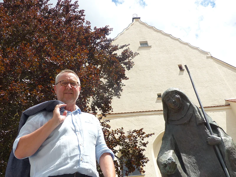 Mann mit Jacke über der Schulter steht neben einer Statue, der Walburga-Statue in Monheim vor einem gelbgestrichenem Gebäude mit Giebeldach. Im Hintergrund ein großer Baum und sonniger Himmel.
