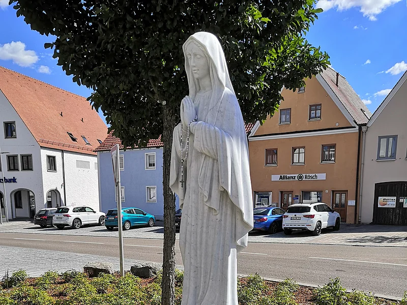 Marienstatue "Maria, Königin des Friedens" Statue einer betenden Frau mit Rosenkranz, die Marienstatue auf einem kleinen Podest, umgeben von Blumen, im Hintergrund eine Straße und ein Parkplatz mit Gebäuden.