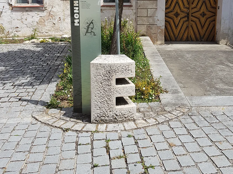 Steinskulptur des Buchstabens "E", dahinter ein Informationsschild und ein Baum mit einem dahinter liegendem Beet. Umgeben von einem gepflastertem Platz, mit einem historischen Gebäude im Hintergrund.
