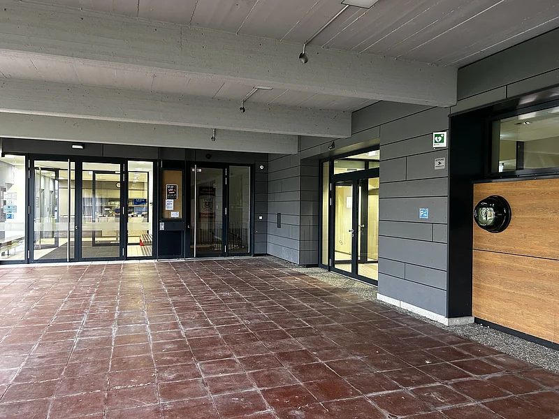 Eingangsbereich mit braunem Fliesenboden, grauen Wänden, mehreren Glastüren und einem Defibrillator an der Wand. Standort Defibrillator Schulsporthalle Richtung Jurabad