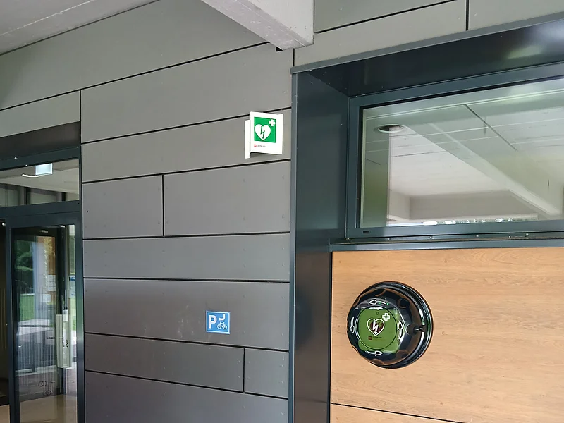 Außenwand mit grauen und holzfarbenen Platten, Defibrillator und Fahrradparkplatzschild neben Glastür. Standort Defibrillator Monheim Schulsporthalle in Richtung Jurabad