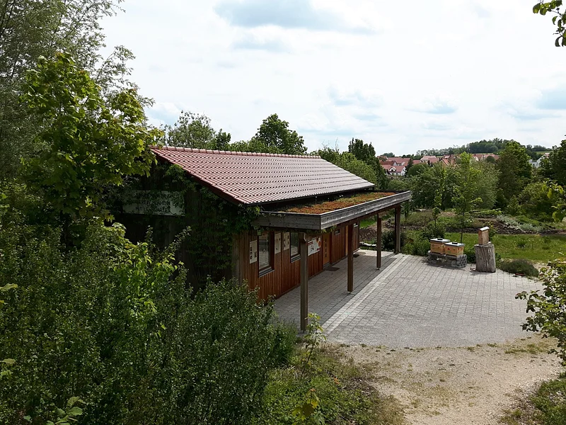Lehrbienenstand mit Bienenweide Monheim Holzgebäude mit rotem Dach und begrüntem Vordach, umgeben von Bäumen und einem gepflasterten Vorplatz, bei bewölktem Himmel.