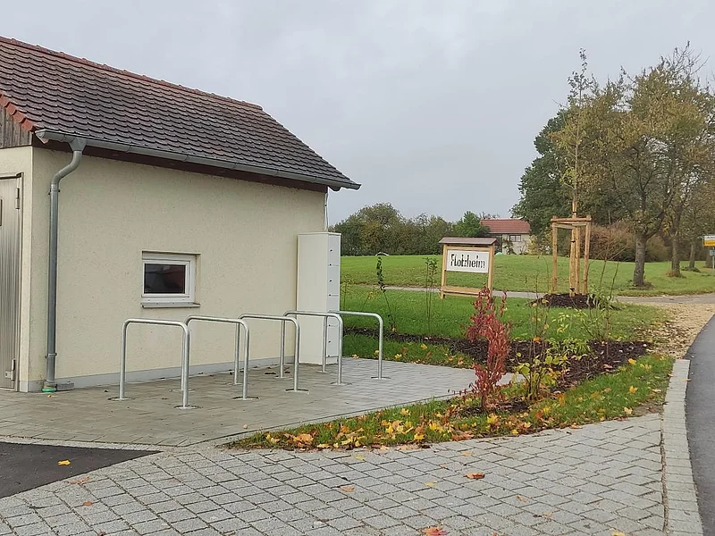 Kleines Gebäude mit Metalltür und kleinem Fenster, sowie fünf Fahrradständern auf gepflastertem Platz neben einer Straße und einem Schild mit Ortsnamen Flotzheim, unter bewölktem Himmel.