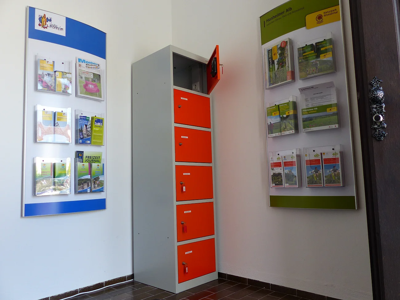 Indoor-Ladeschrank für eBike-Akkus und Infomaterial im Vorraum der Tourist-Information Monheim Ein orange-grauer Indoor-Ladeschrank für eBike-Akkus mit insgesamt sechs Fächern und einem offenen Fach steht zwischen zwei Wanddisplays mit Broschüren in dem Vorraum der Touristinformation Monheim.