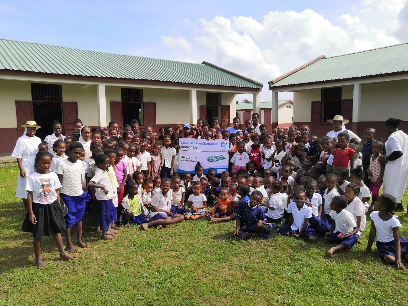 De Lumina unterstützt Schulen in Afrika Große Gruppe von dunkelhäutigen Kindern und Erwachsenen vor Schulgebäuden mit grünem Dach auf einer Wiese stehend und sitzend.
